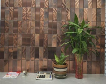 May include: A copper-colored tile wall with a vertical rectangular pattern. A green plant in a clear vase and a smaller potted plant sit on a gray surface. A calculator, stapler, and a magazine are also on the surface.