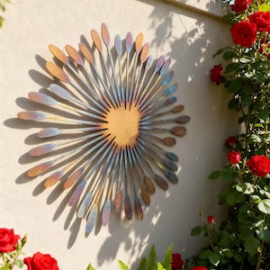 May include: A decorative metal wall sculpture resembling a sunburst, mounted on a wall. The artwork has a central wooden disc, with radiating metal elements in shades of blue, gold, and purple. The wall is surrounded by red roses and green ferns, creating a garden-like setting.