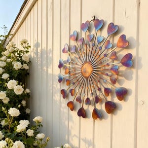 May include: A decorative metal wall hanging with a sunburst design and heart-shaped elements. The hearts display a gradient of colours, including blue, purple, and orange. It's mounted on a white wooden fence, with white roses in the background.