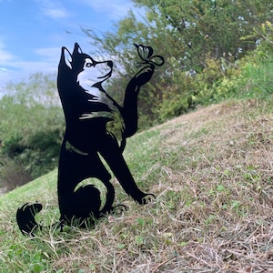 May include: A black metal silhouette of a husky dog sitting in grass, gazing up at a butterfly perched on its paw. The dog has white accents on its face and chest, with a curved tail. The background features green foliage and a blue sky.
