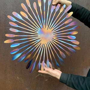 May include: A large, iridescent metal wall art piece. The artwork features a central heart shape with radiating petal-like elements. The colours shift from gold to blue and purple. The piece is held up by two hands.