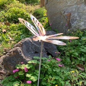 Könnte beinhalten: Ein Gartenstecker aus Metall in Form einer Libelle mit schillernden Flügeln und einem schlanken Stab. Die Libelle befindet sich inmitten von grünem Laub, Felsen und kleinen rosa Blüten und bildet einen dekorativen Akzent im Freien.
