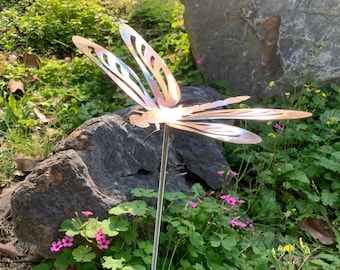 Pode incluir: Uma estaca de jardim de metal em forma de libélula com asas iridescentes e um poste fino. A libélula está posicionada em meio a folhagens verdes, rochas e pequenas flores cor-de-rosa, criando um acento decorativo ao ar livre.