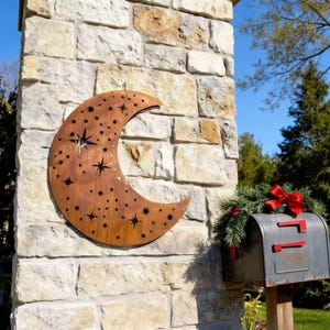 May include: A rustic, crescent moon-shaped metal wall hanging with star cutouts. The brown metal moon is mounted on a stone wall. A mailbox with a red bow and greenery is to the right.