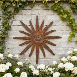 Könnte beinhalten: Ein rostfarbenes Metall-Wandkunstwerk in Form einer Sonne mit einem spiralförmigen Zentrum und spitzen Strahlen. Die Sonne ist in einem warmen, rostbraunen Farbton gehalten und an einer hellgrauen Backsteinmauer befestigt. Weiße Rosen und grüne Weinreben umrahmen das Kunstwerk.