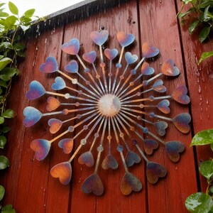 May include: A decorative metal wall hanging with a sunburst design and heart-shaped details. The hearts display a gradient of blue, orange, and purple. It's mounted on a red wooden fence, with green foliage visible in the background.