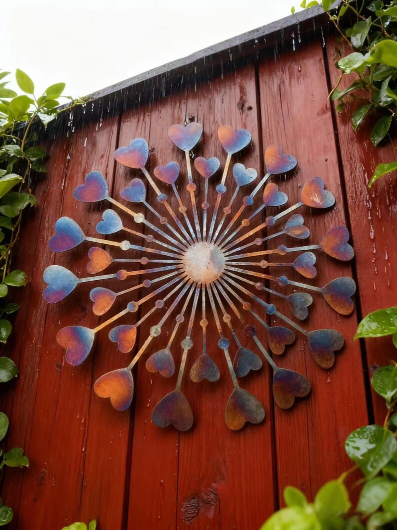 May include: A decorative metal wall hanging with a sunburst design and heart-shaped details. The hearts display a gradient of blue, orange, and purple. It's mounted on a red wooden fence, with green foliage visible in the background.