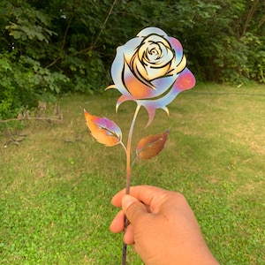 May include: A metal rose garden stake with a colorful, iridescent finish. The flower has a detailed design with a gradient of colors, including blue, orange, and purple. The leaves are also colored with a similar iridescent effect.