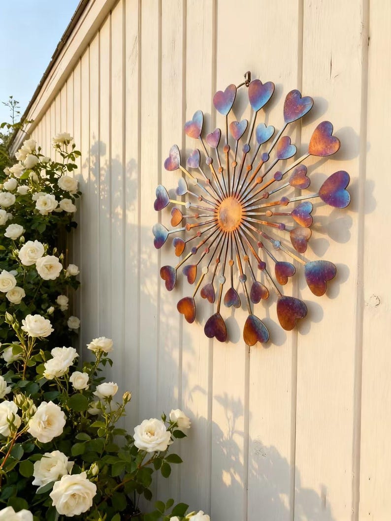 May include: A decorative metal wall hanging with a sunburst design and heart-shaped elements. The hearts display a gradient of colours, including blue, purple, and orange. It's mounted on a white wooden fence, with white roses in the background.