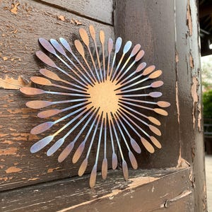 May include: A metal sunburst wall decoration. The design has a central circle with radiating lines, each ending in a spoon shape. The metal has bronze, blue, and gold hues. It is mounted on a weathered wooden door.