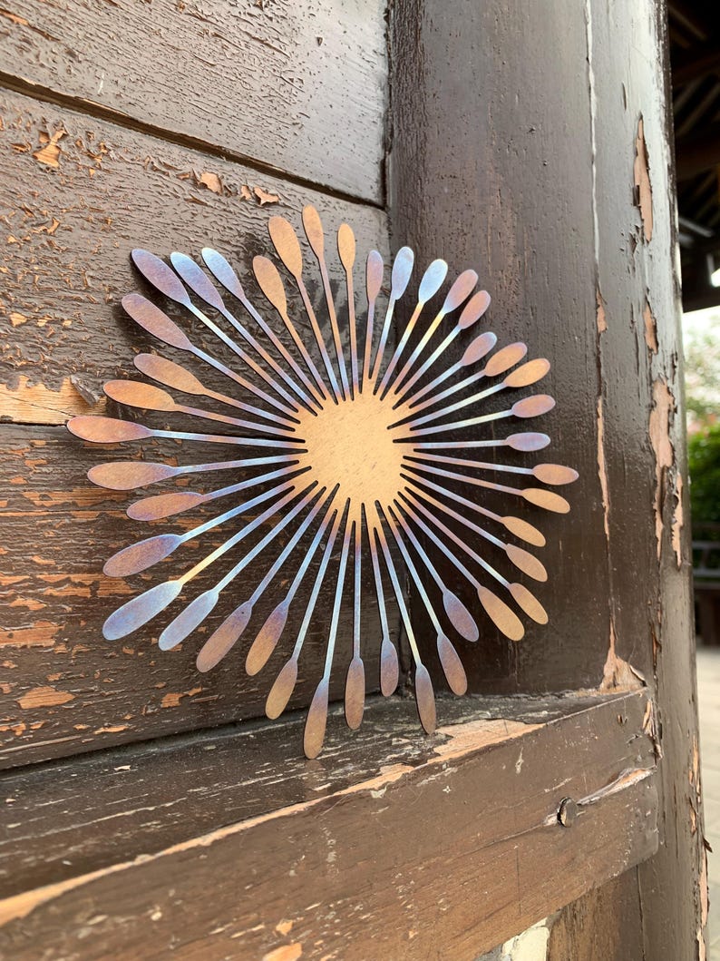 May include: A metal sunburst wall decoration. The design has a central circle with radiating lines, each ending in a spoon shape. The metal has bronze, blue, and gold hues. It is mounted on a weathered wooden door.