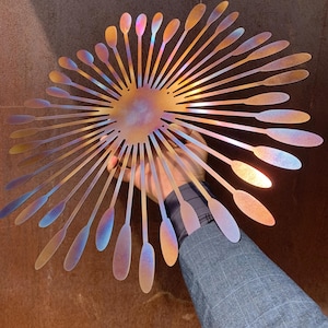 May include: A decorative metal wall art piece in a sunburst design. It features a central circular element with radiating, elongated petal-like shapes. The metal has an iridescent finish, displaying gold, blue, and purple hues.