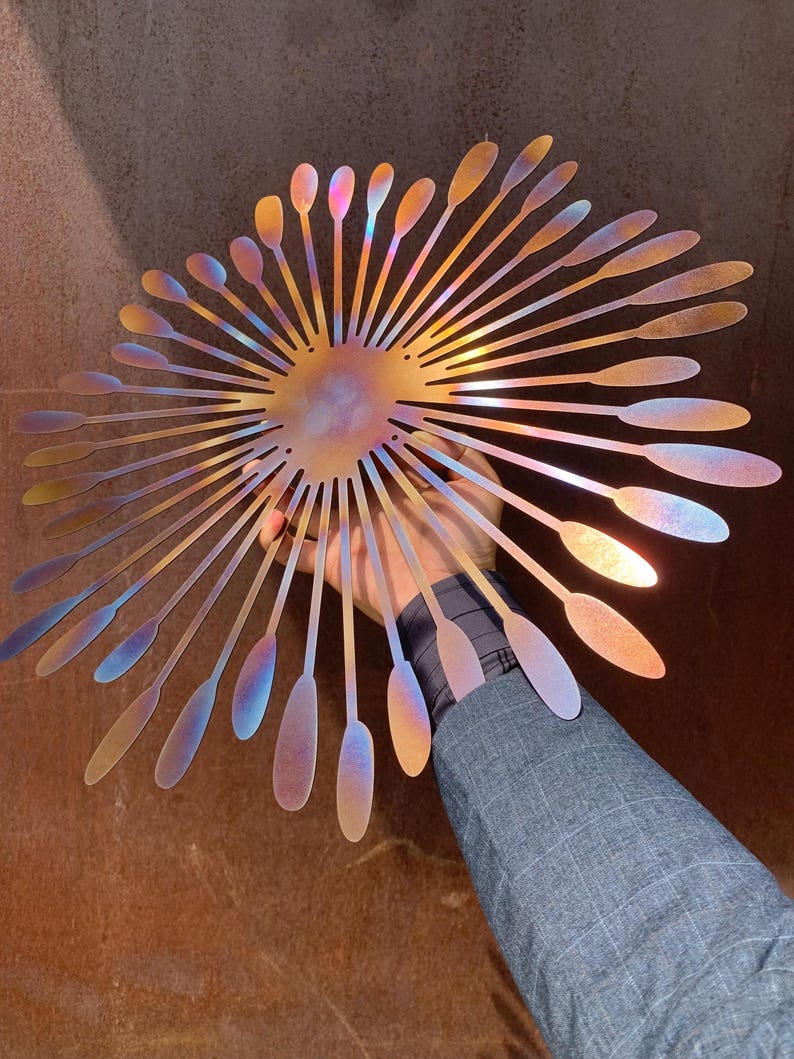 May include: A decorative metal wall art piece in a sunburst design. It features a central circular element with radiating, elongated petal-like shapes. The metal has an iridescent finish, displaying gold, blue, and purple hues.
