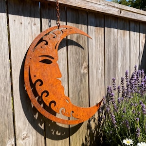 May include: A rusty metal crescent moon with a face design hangs from a chain on a wooden fence. The moon has decorative cutouts and is positioned near a lavender bush and daisies, creating a garden-themed decoration.
