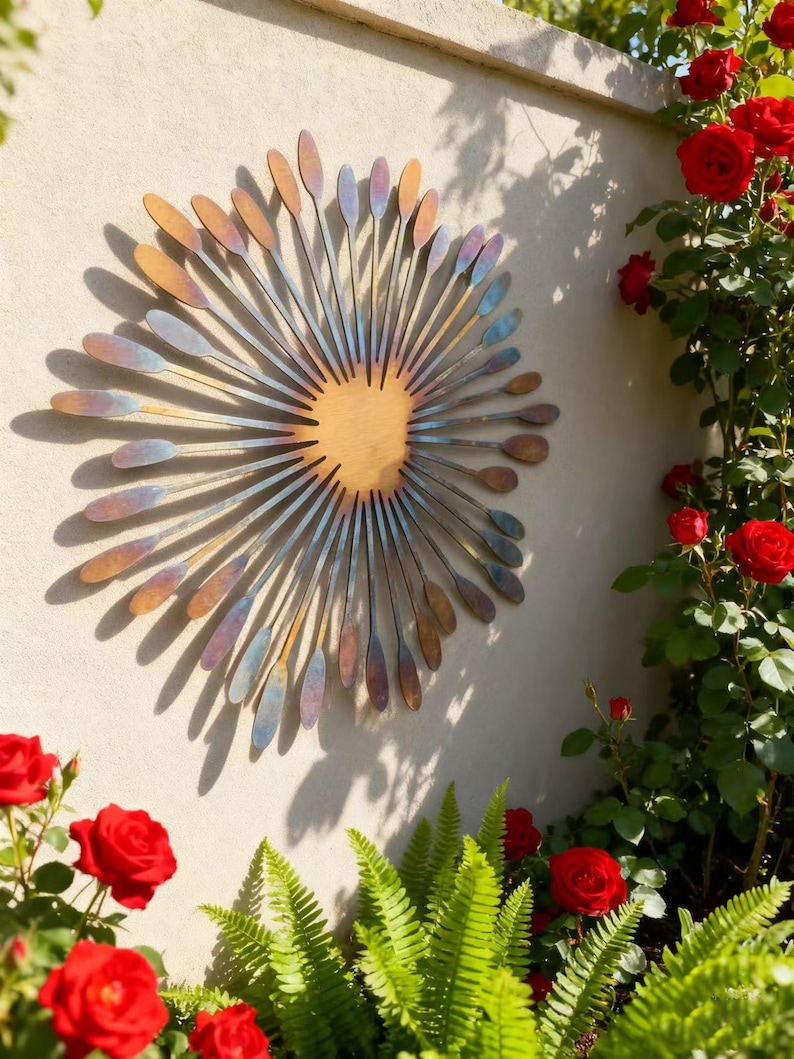 May include: A decorative metal wall sculpture resembling a sunburst, mounted on a wall. The artwork has a central wooden disc, with radiating metal elements in shades of blue, gold, and purple. The wall is surrounded by red roses and green ferns, creating a garden-like setting.