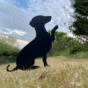 May include: A black metal silhouette of a dog in a sitting position, with one paw raised. The dog is positioned in a grassy area with trees and a cloudy sky in the background. The dog's tail is curled.