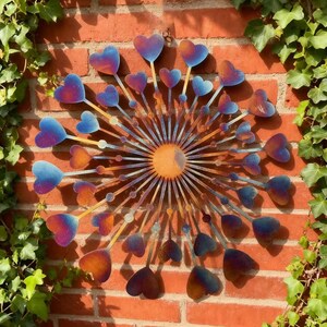 May include: Metal wall art with a sunburst design and heart-shaped elements. The hearts are coloured blue to orange, set against a red brick wall with green ivy, creating a vibrant display.