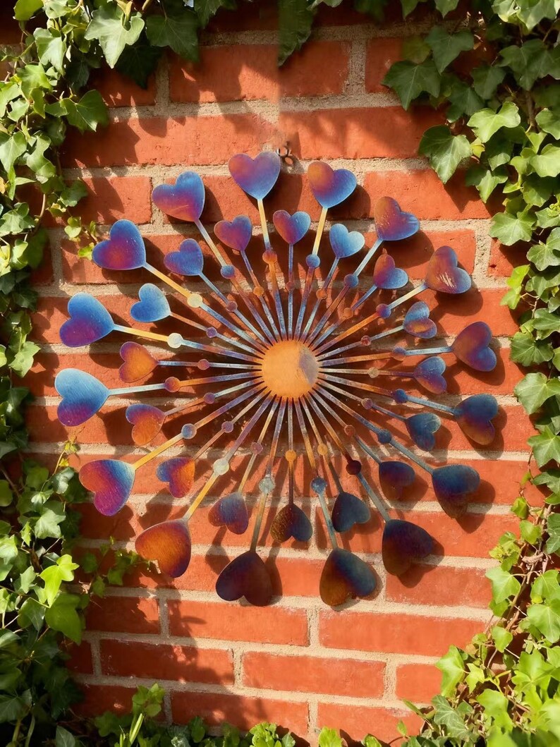 May include: Metal wall art with a sunburst design and heart-shaped elements. The hearts are coloured blue to orange, set against a red brick wall with green ivy, creating a vibrant display.