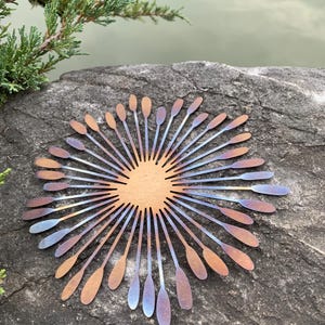 May include: A metal sunburst sculpture with a central circular design and radiating petal-like elements. The petals transition in colour from copper to blue and purple. The sculpture is displayed outdoors on a rock.