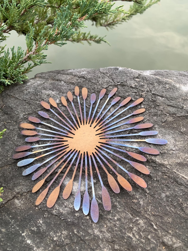 May include: A metal sunburst sculpture with a central circular design and radiating petal-like elements. The petals transition in colour from copper to blue and purple. The sculpture is displayed outdoors on a rock.