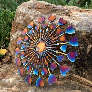 May include: A metal garden ornament with a sunburst design and heart-shaped petals. The piece displays a gradient of colours, including orange, blue, and purple. It is mounted on a large, textured rock, creating a decorative outdoor feature.