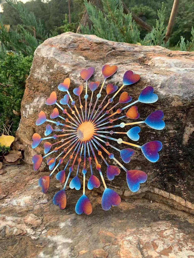 May include: A metal garden ornament with a sunburst design and heart-shaped petals. The piece displays a gradient of colours, including orange, blue, and purple. It is mounted on a large, textured rock, creating a decorative outdoor feature.