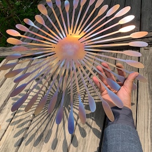 May include: A decorative metal wall art piece in a sunburst design. It features a central circular element with radiating, elongated petal-like shapes. The metal has a colour-shifting finish, displaying hues of gold, purple, and blue.
