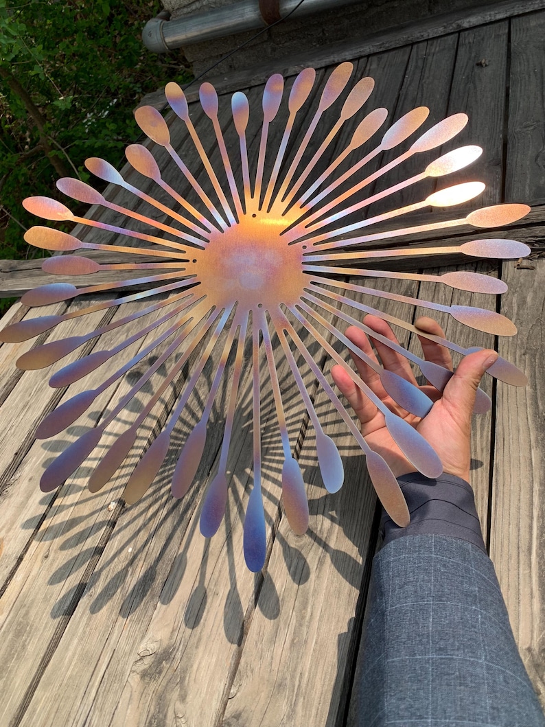 May include: A decorative metal wall art piece in a sunburst design. It features a central circular element with radiating, elongated petal-like shapes. The metal has a colour-shifting finish, displaying hues of gold, purple, and blue.