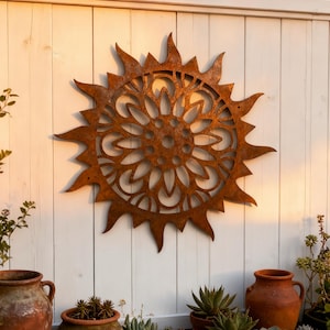 May include: A rusty metal sunburst wall art with a detailed floral center is mounted on a white wooden fence. The sunburst has pointed rays and a circular, openwork design. The scene includes terracotta pots with succulents and other plants.