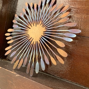 May include: Metal wall art depicting a sunburst design. The central area is a warm gold hue, with radiating lines that shift from bronze to iridescent blue and purple. It's mounted on a wooden surface.