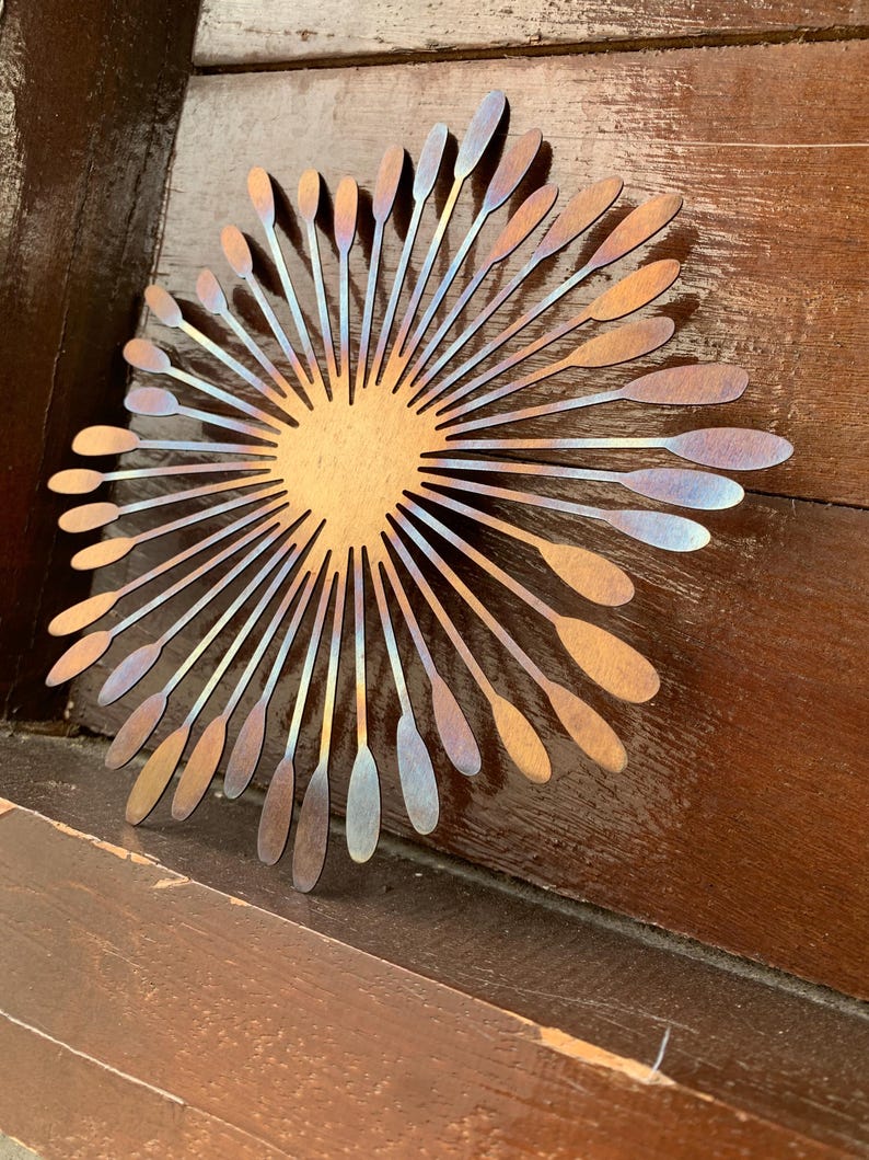 May include: Metal wall art depicting a sunburst design. The central area is a warm gold hue, with radiating lines that shift from bronze to iridescent blue and purple. It's mounted on a wooden surface.