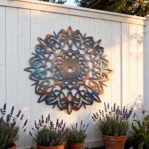 Op de afbeelding: Een groot, cirkelvormig metalen wandkunstwerk met een gedetailleerd, opengewerkt ontwerp in tinten blauw en brons. Het kunstwerk is gemonteerd op een witte houten schutting, met potlavendelplanten op de voorgrond, wat een tuin thema creëert.