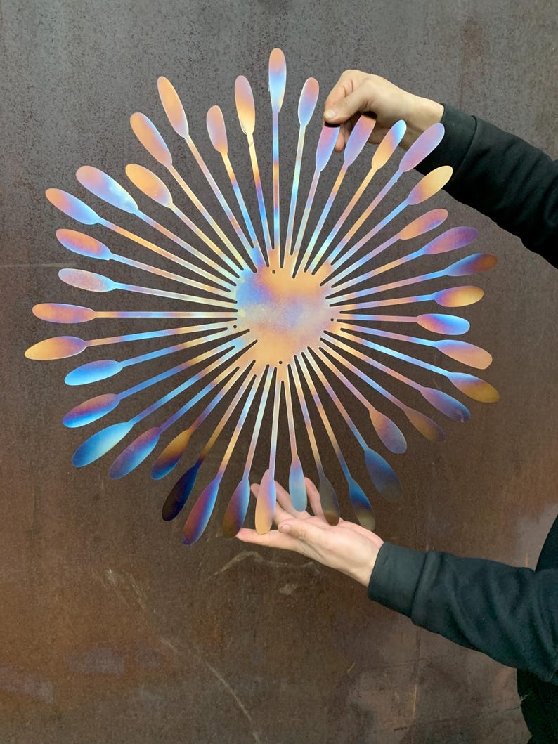 May include: A large, iridescent metal wall art piece. The artwork features a central heart shape with radiating petal-like elements. The colours shift from gold to blue and purple. The piece is held up by two hands.