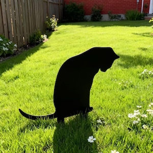 Könnte beinhalten: Schwarze Metall-Katzen-Silhouette als Gartenstecker. Die Katze sitzt mit ausgestrecktem Schwanz. Der Stecker befindet sich in einem grasbewachsenen Garten mit weißen Blumen und einem Holzzaun im Hintergrund.