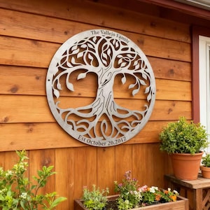 Puede incluir: Una pieza de arte mural de metal circular con un árbol genealógico, con el texto "The Vallejo Family" y "Est October 20, 2012". El diseño del árbol presenta un tronco, ramas y hojas detallados. Está montado en una pared de madera.