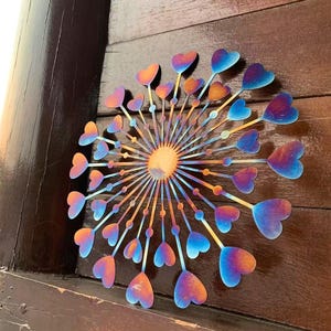 May include: A metal wall art piece featuring a dandelion design with heart-shaped seed heads. The piece has a gradient of colours, including orange, blue, and purple. The artwork is mounted on a wooden surface.