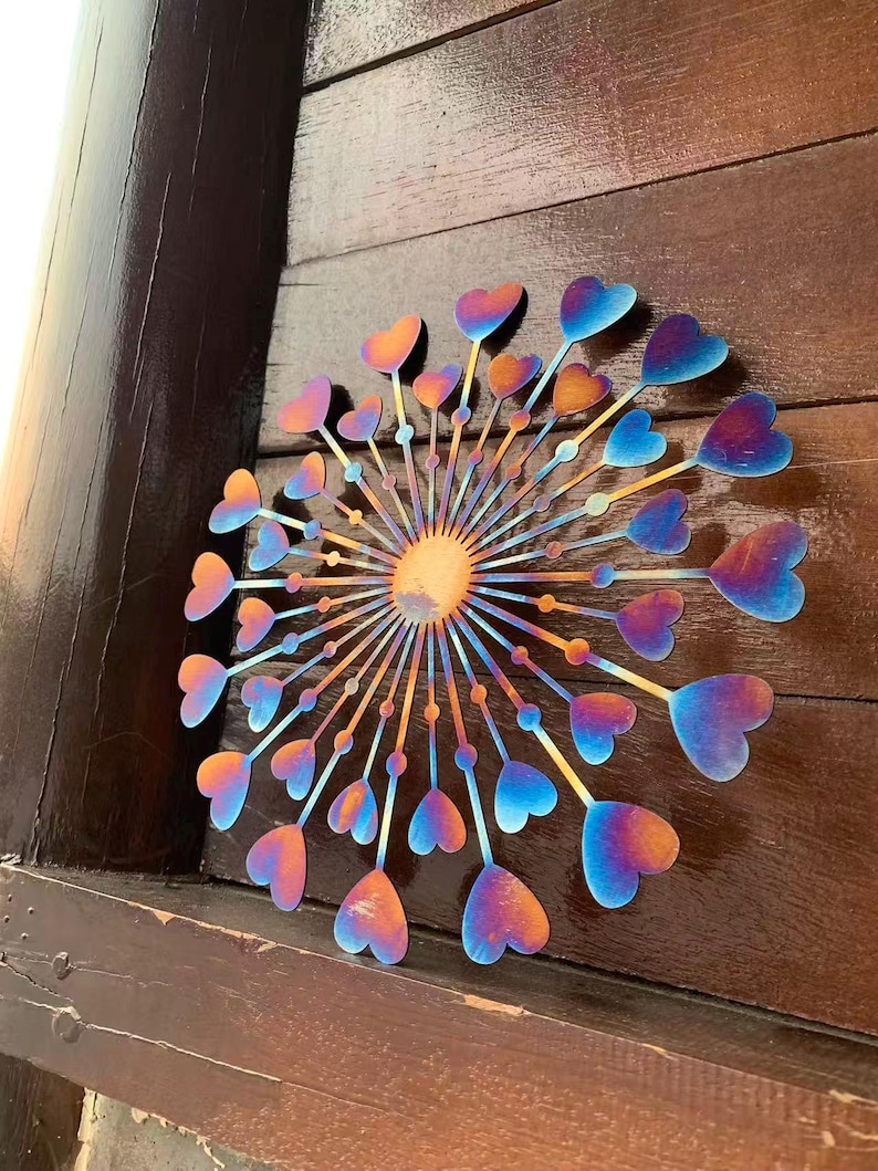 May include: A metal wall art piece featuring a dandelion design with heart-shaped seed heads. The piece has a gradient of colours, including orange, blue, and purple. The artwork is mounted on a wooden surface.