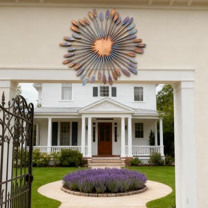May include: A large metal sunburst sculpture with a copper centre and radiating blades in shades of blue, purple, and copper. The artwork is mounted on a beige wall above a white house with a brown door.