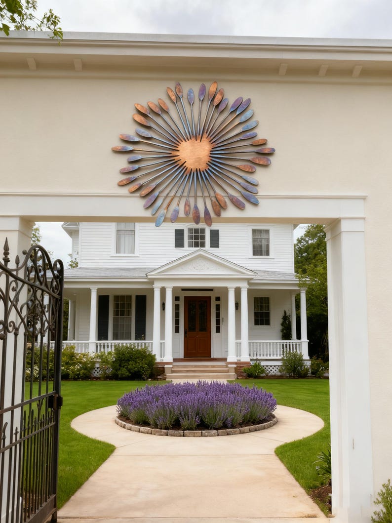 May include: A large metal sunburst sculpture with a copper centre and radiating blades in shades of blue, purple, and copper. The artwork is mounted on a beige wall above a white house with a brown door.