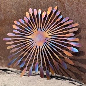 May include: Metal wall art in a sunburst design. The artwork features a central circular element with radiating lines ending in teardrop shapes. The metal has a colour gradient of bronze, gold, blue, and purple.