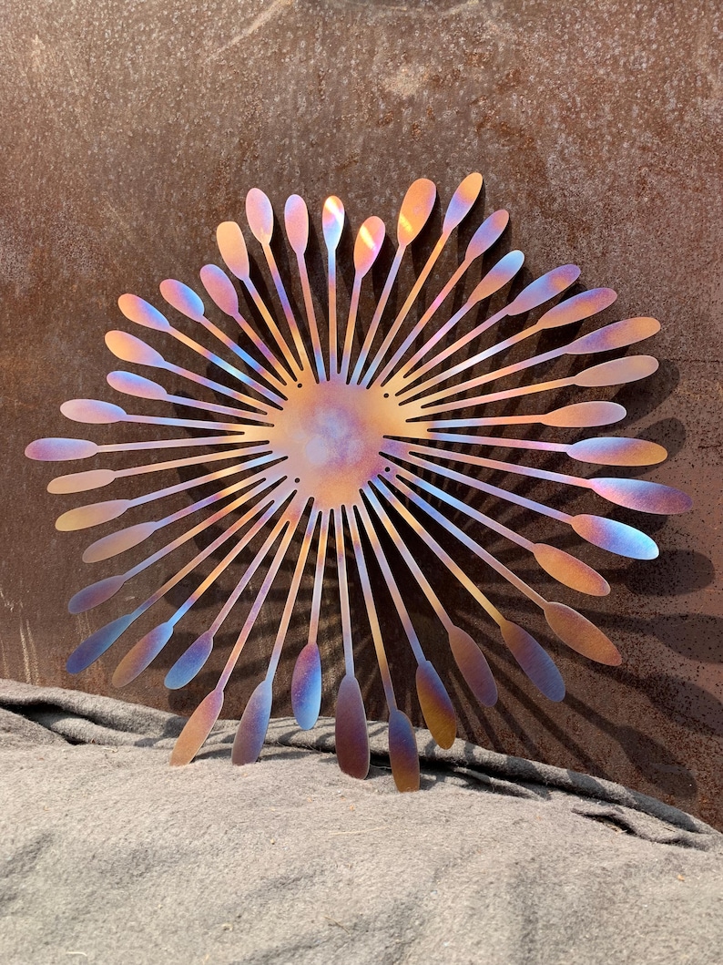 May include: Metal wall art in a sunburst design. The artwork features a central circular element with radiating lines ending in teardrop shapes. The metal has a colour gradient of bronze, gold, blue, and purple.
