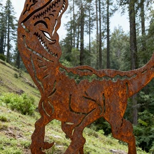 Puede incluir: Escultura de metal marrón oxidado de un Tyrannosaurus Rex sonriente, con detalles recortados. La escultura está sobre una colina cubierta de hierba, con árboles altos de fondo, creando una decoración exterior caprichosa.