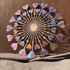 May include: A circular metal wall art piece depicting a dandelion with heart-shaped seed heads. The piece has a gradient of colours, including pink, blue, and gold. The artwork has a central sun-like element.