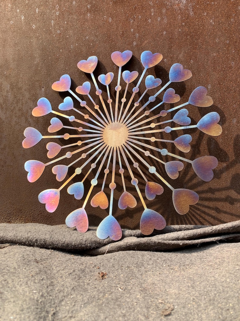 May include: A circular metal wall art piece depicting a dandelion with heart-shaped seed heads. The piece has a gradient of colours, including pink, blue, and gold. The artwork has a central sun-like element.