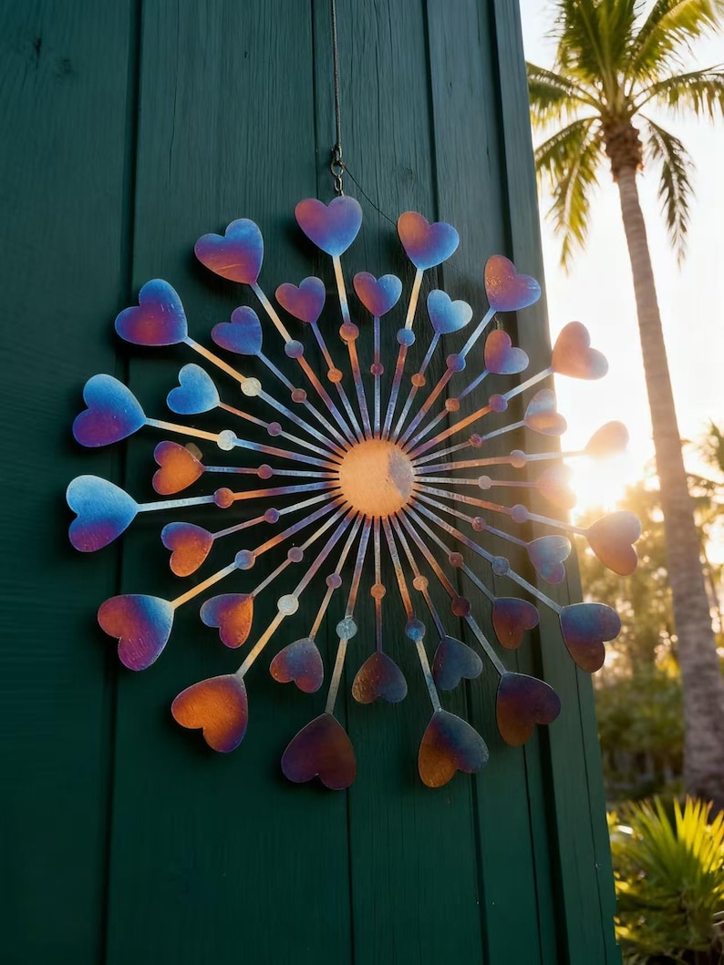 May include: A decorative metal wall hanging featuring a heart design. The hearts are coloured in shades of blue, purple, and orange, emanating from a central sun-like form. It is suspended by a wire and hangs against a green wooden surface.
