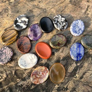 May include: A collection of oval-shaped gemstones in various colors and patterns, including tiger's eye, amethyst, and jasper. The stones are arranged on a textured, brown surface, showcasing their natural beauty and unique textures.