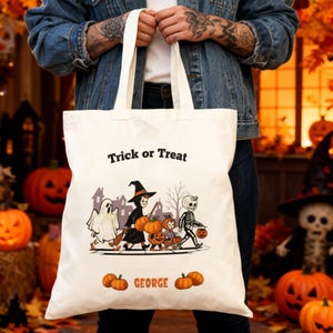 Puede incluir: Un bolso tote color crema con las palabras "Trick or Treat" impresas en la parte superior. El bolso presenta una ilustración de Halloween de un fantasma, una bruja y un esqueleto pidiendo dulces. El nombre "GEORGE" está impreso en la parte inferior, con calabazas a cada lado.