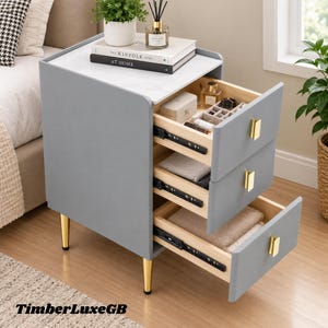 May include: A gray bedside table with a white marble-patterned top, three drawers with gold rectangular handles, and four gold tapered legs. The top holds a plant and a book titled The Kinfolk Home. The drawers contain various small items.