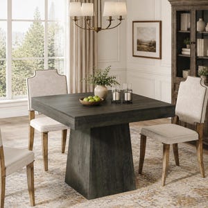 May include: A square dining table with a dark gray wood grain finish and a wide pedestal base, accompanied by four upholstered chairs featuring light beige fabric, nailhead trim, and wooden legs, arranged in a bright, neutral-toned room.