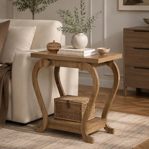 May include: A light brown wooden side table with a rectangular top and a lower shelf. The table features four curved legs. On the top surface sits a white vase with greenery, two stacked books, and a small brown lidded container. A woven basket sits on the lower shelf.